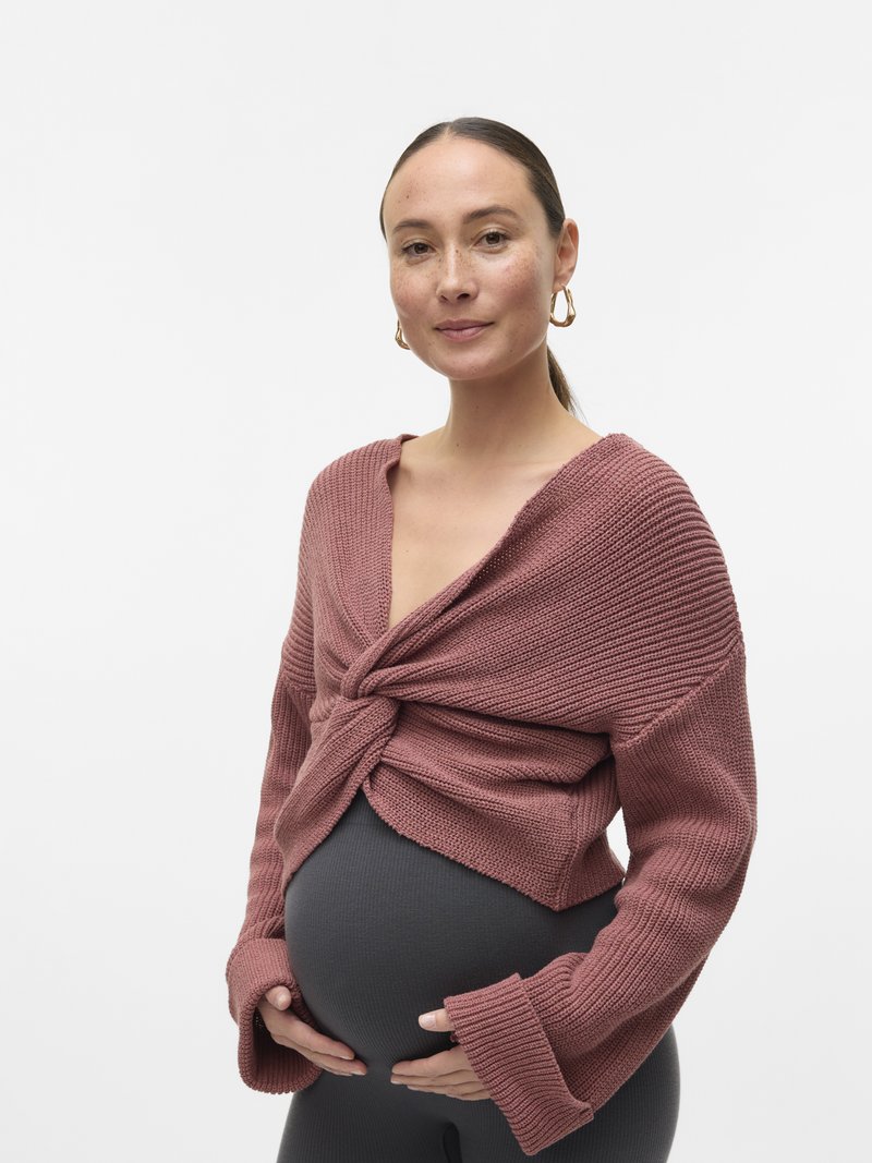 Femme enceinte aux longs cheveux attachés, portant un pull mauve en tricot sur un legging noir, tenant doucement son ventre et souriant doucement.