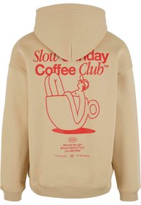 Beige hættetrøje med et stort rødt grafisk motiv, der viser en person liggende i en kaffekop. Teksten inkluderer "Slow Sunday Coffee Club" og yderligere detaljer.