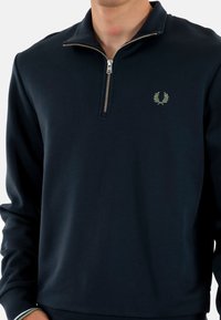 Sudadera con cremallera en azul marino de tejido suave. Presenta una cremallera plateada, un cuello alto, puños acanalados y un pequeño logo verde en el pecho.