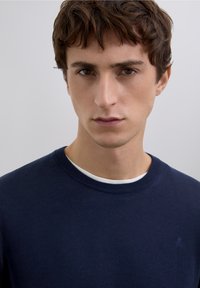 Mörkblå crewneck-tröja i mjukt tyg med en strukturerad stickning och ett vitt innerfoder i kragen. Liten logga på bröstet.