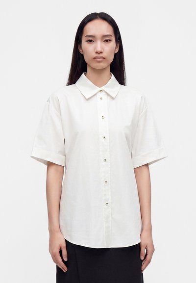 Femme aux longs cheveux noirs portant une chemise blanche à manches courtes boutonnée et une jupe noire sur un fond blanc uni.