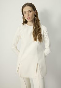 White long-sleeve top with side slits, made of smooth fabric. Simple round neckline and fitted design. Model wearing large, curved earrings.