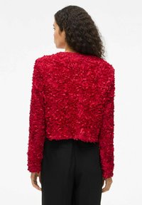 Veste courte à texture florale rouge avec des manches longues, présentant des détails en couches en forme de pétales et un col rond, associée à un pantalon noir.