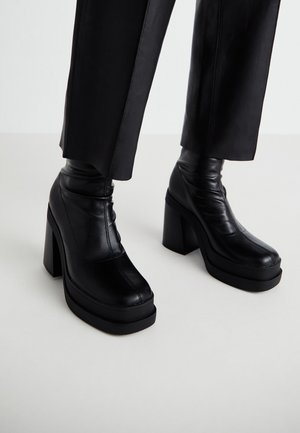 Botas de plataformas negras de cuero con un alto tacón de bloque y una textura suave. Presentan detalles de costura y un diseño de parte superior ajustada.