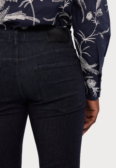 Jeans de mezclilla oscura con un diseño ajustado, que cuentan con bolsillos traseros y un logotipo en la cintura de cuero negro. Combinados con una camisa de patrón floral.