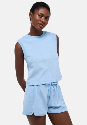 Femme portant un haut sans manches bleu clair et un short assorti avec cordon, debout avec les bras dans le dos sur fond blanc.