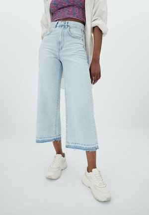 Person wearing light blue wide-leg cropped jeans, white chunky sneakers, multicolored crop top, and white open shirt standing on white background.