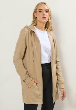 Femme portant une veste à capuche beige zippée par-dessus un t-shirt blanc et un pantalon noir, debout devant un fond clair uni.