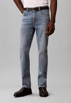 Homme portant un jean bleu clair, une ceinture noire, une chemise blanche, des chaussures en cuir noires et un bracelet en argent, debout sur un fond uni.