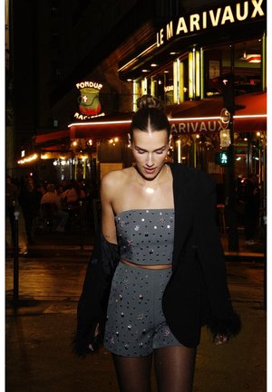 Jeune femme en top tube à paillettes grises et en shorts avec une veste noire passe devant un café illuminé par la nuit dans un cadre urbain.