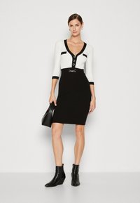 Robe ajustée noir et blanc avec des manches trois-quarts, décolleté en V, détails de boutons et une ceinture à la taille. Portée avec des bottines noires.