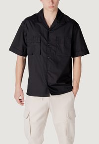 Camisa negra de manga corta con botones, cuello puntiagudo, dos bolsillos en el pecho y una tela suave y ligera. Combinada con pantalones cargo color crema.