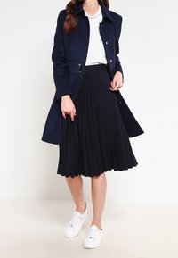 Manteau en mélange de laine marine avec col et fermeture à bouton, associé à une jupe plissée noire et des baskets blanches. Tissu lisse, design mi-long.