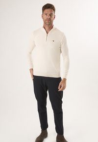 Cremfarbener Pullover mit Reißverschluss am Hals, langen Ärmeln, gerippten Bündchen und einem kleinen braunen Logo auf der Brust, kombiniert mit dunklen Hosen und braunen Stiefeln.
