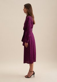 Abito fucsia a maniche lunghe con motivo geometrico nero, vita cintrata e orlo svasato; indossato con sandali neri con tacco alto e cinturini.