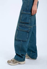 Pantalones cargo de mezclilla azul con grandes bolsillos laterales, piernas anchas y costuras visibles. Llevados con zapatillas deportivas beige.