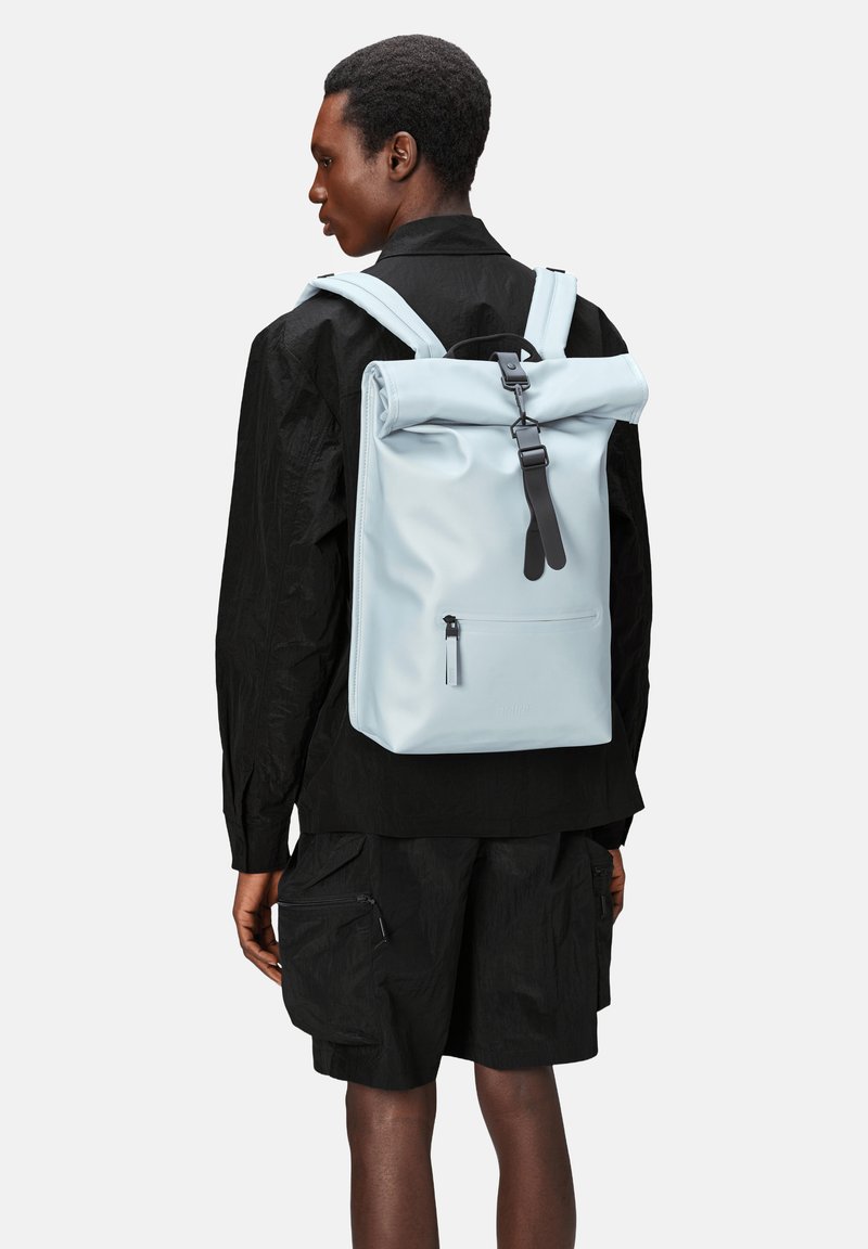 Mochila de color azul claro con tapa enrollable, fabricada en un material suave, que presenta herrajes negros y un bolsillo con cremallera en la parte frontal. Lleva puesta sobre una camiseta negra.