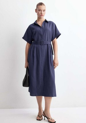 Femme portant une robe midi bleu marine cintrée avec manches courtes, tenant un sac à main noir tissé et portant des sandales noires à talons.