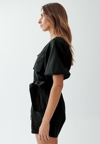Vestido corto negro de manga corta con forma ajustada, que cuenta con un cinturón en la cintura, bolsillos laterales y detalles de volantes en capas en el bodice.