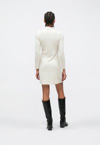 Rochie tricotată crem cu mâneci lungi și croială strâmtă. Purtată cu cizme negre înalte. Stil minimalist, textură netedă și un model subtil.