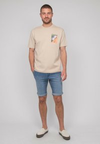 T-shirt en coton beige avec un motif de palmier sur une patch. Assorti à un short en denim roulé et des chaussures légères à enfiler.