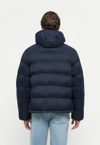Man in een marineblauwe gewatteerde jas met capuchon, van achteren gezien, gecombineerd met lichtblauwe denim jeans, tegen een eenvoudige witte achtergrond.