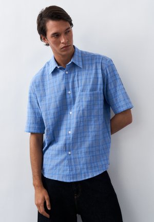 Mand iført blå kortærmet skjorte med ternet mønster, stående mod en hvid væg, kigger til sin venstre side.