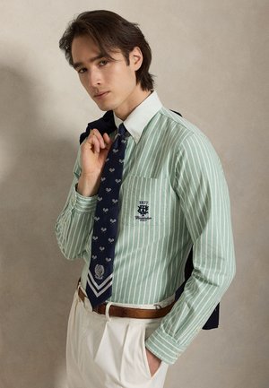 Tânăr purtând o cămașă verde cu dungi, cu logo-ul Wimbledon Polo, cravată bleumarin cu motive de rachete de tenis, pantaloni albi și ținând o jachetă pe umăr.