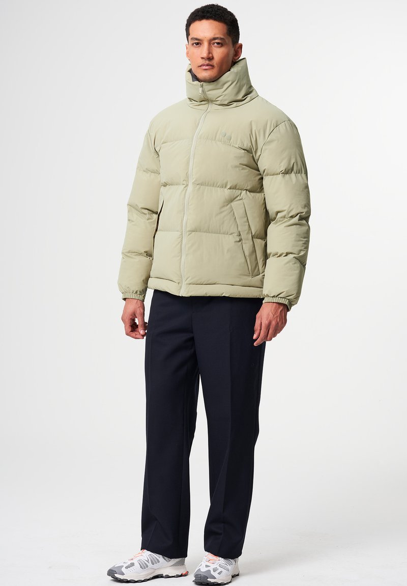 Hellgrüne gepolsterte Jacke mit hohem Kragen, Frontreißverschluss und Seitentaschen, kombiniert mit marineblauen Hosen und grauen Laufschuhen.