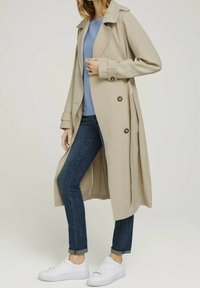 Manteau beige avec deux gros boutons, manches longues et col en V, associé à un haut bleu clair, un jean foncé et des baskets blanches.