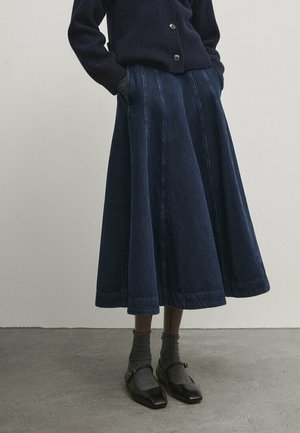 Personne portant une jupe midi en jean bleu foncé coupe A, un cardigan noir boutonné, des chaussettes grises et des chaussures Mary Jane noires à bout carré.