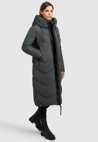 Lang, mørkegrå pufferjakke med chevron-quilting, stor hætte og lynlås foran. Har snoretræk og sidelommer. Midt på læggen længde.