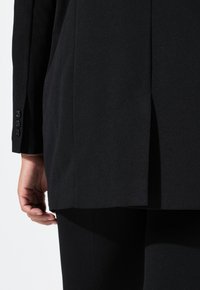 Blazer nero su misura con design strutturato, texture liscia e uno spacco sul retro. I bottoni sui polsini delle maniche sono visibili.