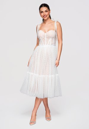 Femme portant une robe midi blanche transparente à pois avec bustier corset, bretelles nouées aux épaules et chaussures transparentes à talons hauts, souriante et posant.