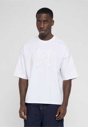 Jonge man draagt een losse witte T-shirt met een gestudeerd abstract ontwerp en donkerblauwe spijkerbroek, staand tegen een effen achtergrond.