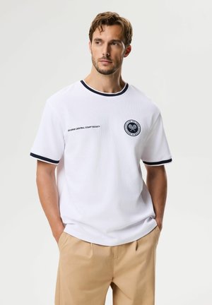 Mężczyzna z krótkimi brązowymi włosami, ubrany w biały t-shirt z krótkim rękawem z czarnym obszyciem i beżowe spodnie, stoi z rękami w kieszeniach.