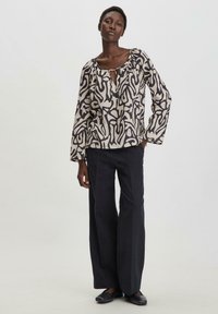 Blusa larga beige con un motivo astratto nero, con scollatura a chiave e maniche lunghe. Abbinata a pantaloni neri a gamba larga.