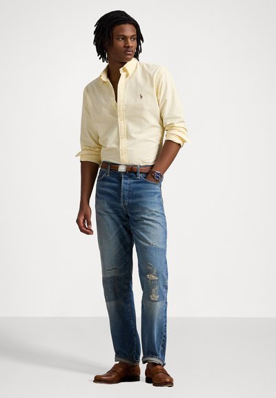 Camisa botão amarela clara, jeans azul desgastados, cinto de couro marrom e mocassins marrons. Apresenta um pequeno logotipo na camisa.