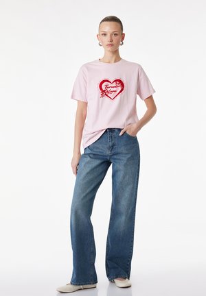 Jeune femme portant un t-shirt rose avec un cœur rouge et le texte « Forever Love », associée à un jean bleu et des chaussures blanches, posant avec une main dans la poche.