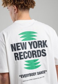 Valkoinen puuvillapaita, jossa on mustaa tekstiä "NEW YORK RECORDS" ja vihreitä graafisia yksityiskohtia, sekä lainaus "EVERYBODY DANCE" alla.