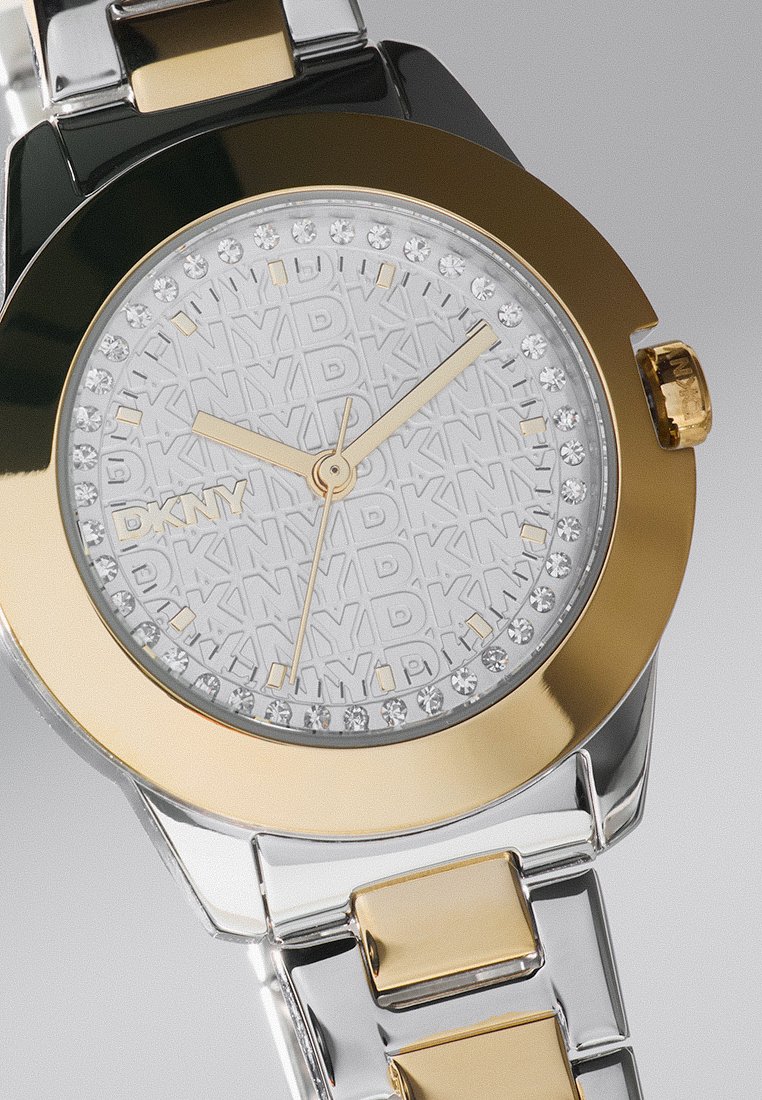 Montre DKNY avec un bracelet métallique argenté et doré, un cadran rond, un cadran en argent texturé, des index d'heure dorés et des accents en cristaux scintillants.