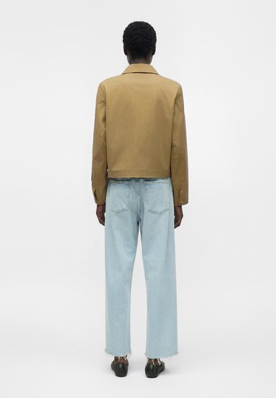 Άτομο που φοράει κοντό μπεζ μπουφάν και ανοιχτό μπλε παντελόνι με φαρδιά πόδια και φθαρμένα άκρα, εμφανίζεται από πίσω σε λευκό φόντο.