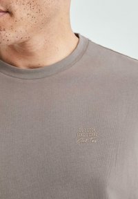 Graues Baumwoll-T-Shirt mit Rundhalsausschnitt, das mit einem hellgoldenen gestickten Text versehen ist: "SEHE ICH AUS, ALS OB ES MICH INTERESSIERT" und "Gute Zeiten" auf der Brust.