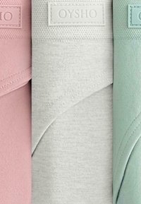 Drie opgerolde yogamatten in roze, grijs en mintgroen. Elke mat heeft een gestructureerde afwerking met een klein, wit "OYSHO" label.