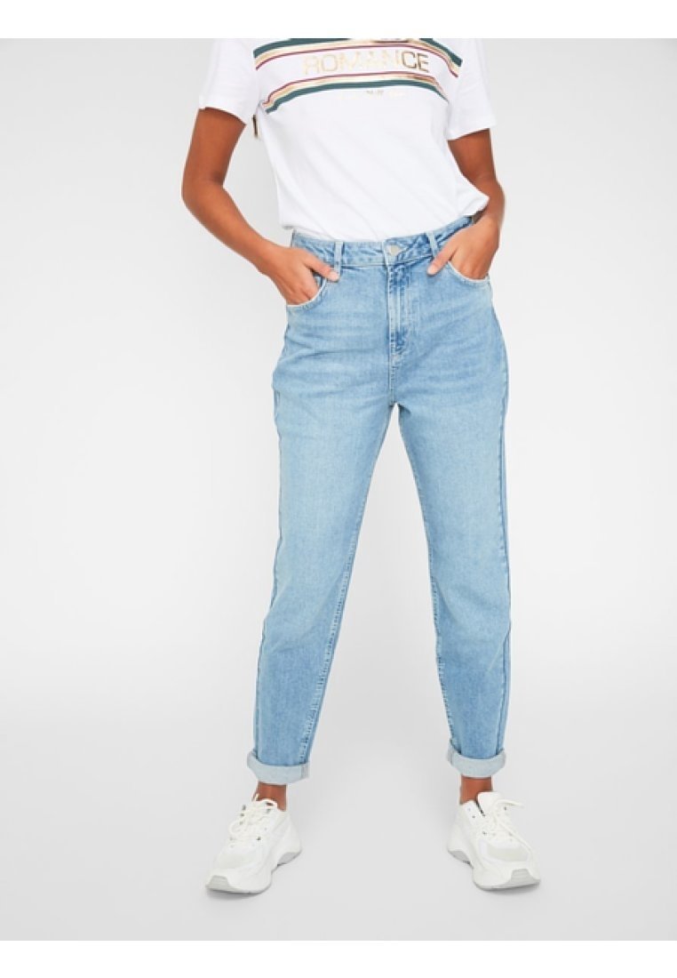 Personne portant un jean taille haute bleu clair roulé aux chevilles, un t-shirt blanc à motif graphique et des baskets blanches, debout les mains dans les poches.