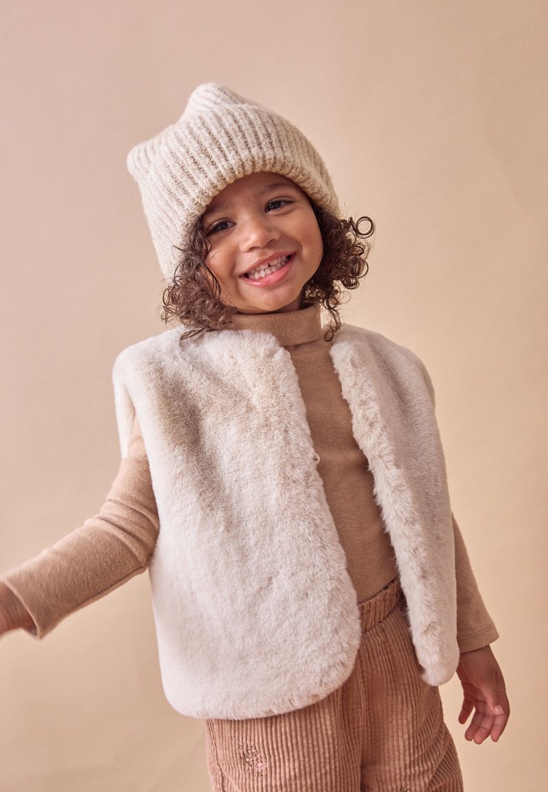 Enfant souriant portant un bonnet en tricot beige, un col roulé marron clair, un gilet en fausse fourrure crème et un pantalon en velours côtelé marron, sur un fond beige.