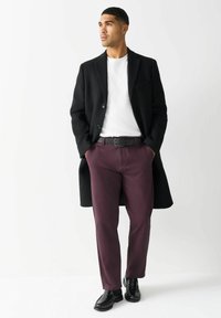 Czarny dopasowany płaszcz, biały T-shirt, bordowe spodnie i błyszczące czarne buty. Guziki na płaszczu, slim fit, gładkie tekstury w całej odzieży.