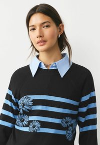 Black sweater with blue stripes and floral embroidery. Features a collared light blue shirt layered underneath. Textured knit fabric.