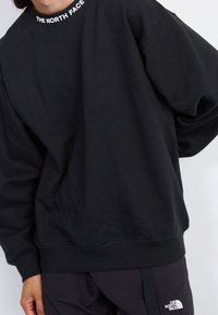 Osoba oblečená v černé mikině s dlouhým rukávem a kalhotách, oba s logem "The North Face" na límci a levé nohavici.