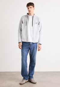 Veste zippée gris clair avec un panneau supérieur texturé, des accents noirs et deux poches avant, assortie à un jeans bleu et des chaussures grises.